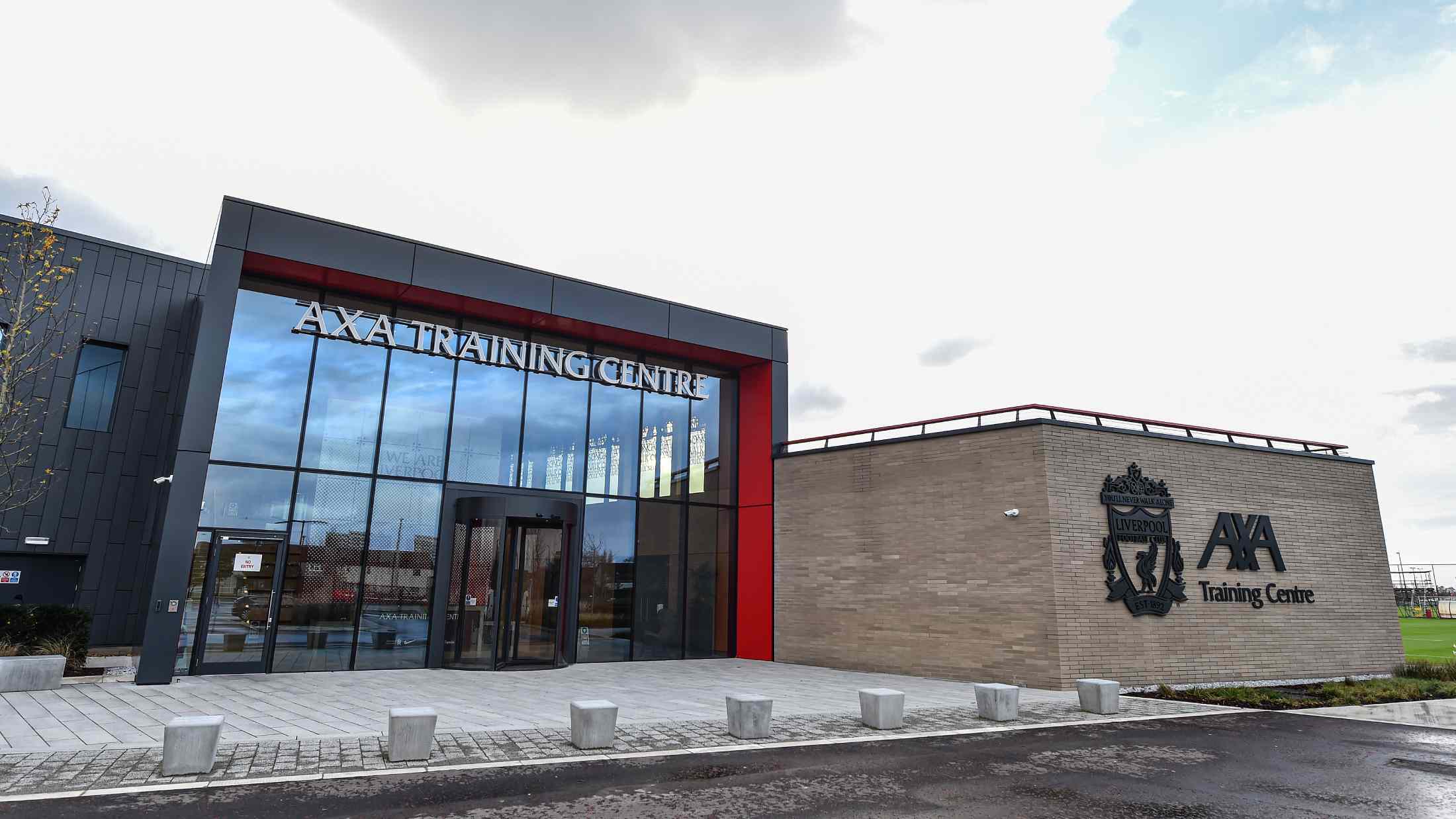 Outside of the Melwood training center for Liverpool FC, a large building with floor to ceiling glass and the liverpool fc logo and the AXA logo on the outside