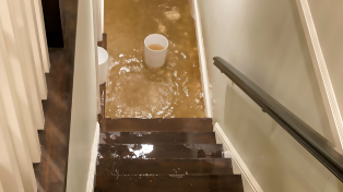 A bucket at the bottom of the stairs in a house flooded with dirty brown water
