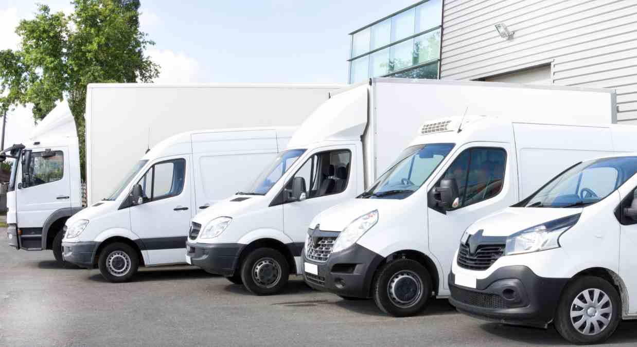 Five white vans lined up outside in a car park outside a warehouse