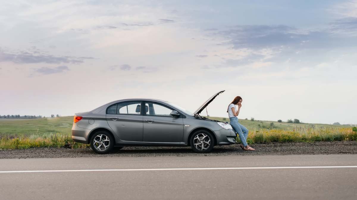 A frustrated young girl stands near a broken-down car at the side of the road