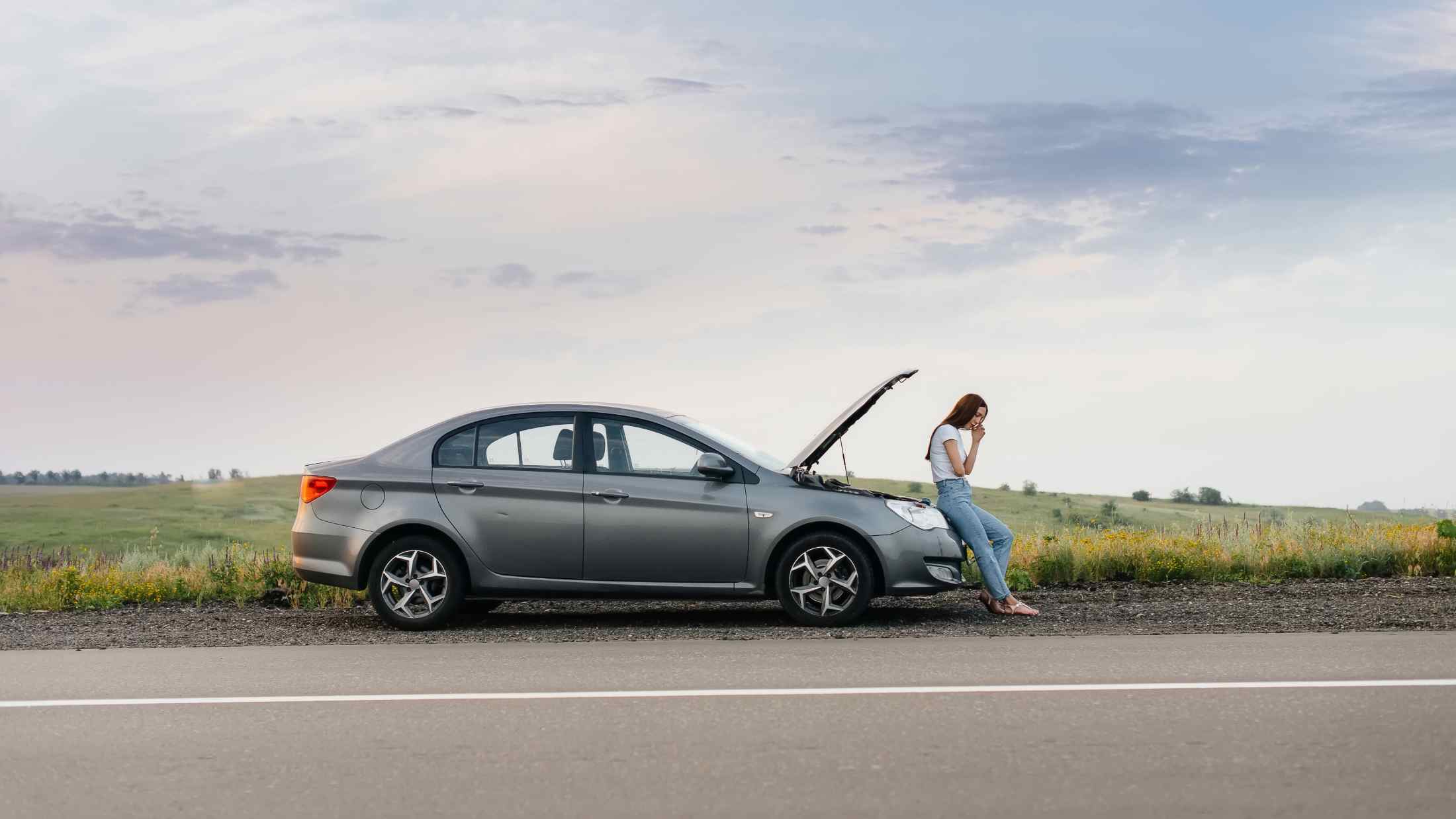 A frustrated young girl stands near a broken-down car at the side of the road