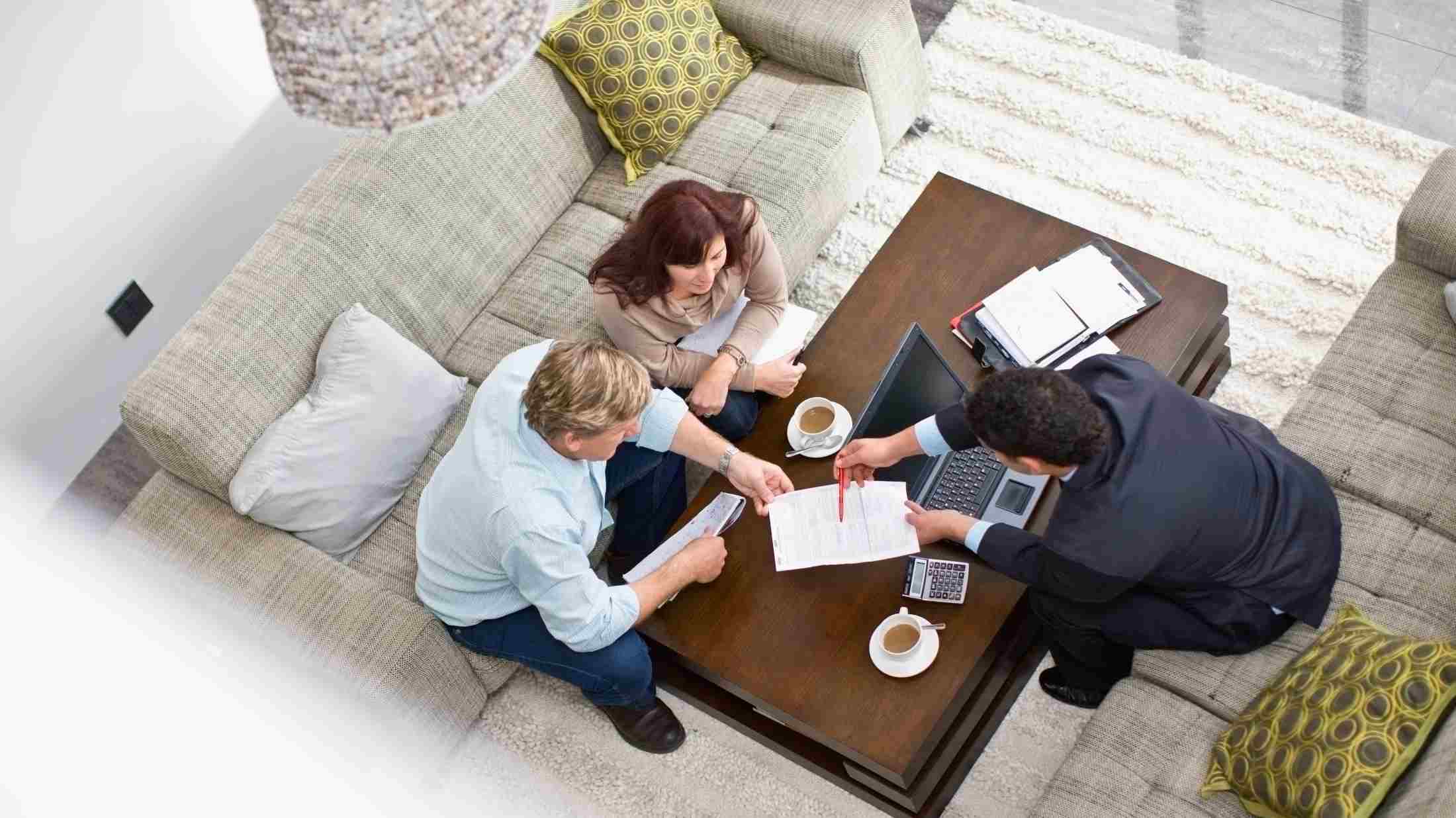 A couple sit together on a sofa reviewing paperwork shown to them by a professional man with a suit who sites on another sofa