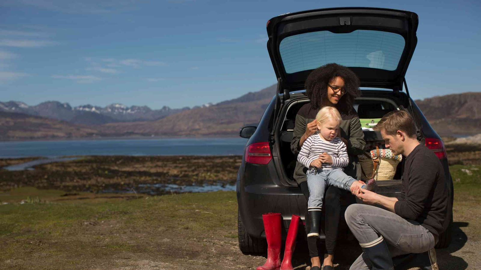 Mum, dad and young child changing shoes in the back of their car in the countryside with mountain scenery behind them