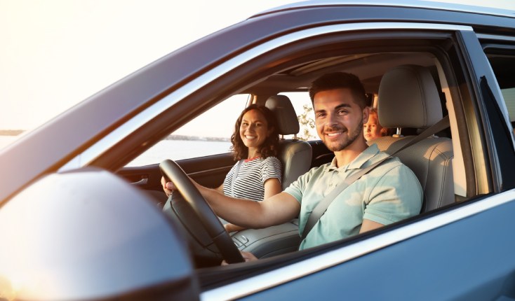 Photo of a family of three sat in a car taken from the drivers side window, the parents are smiling towards the camera whilst the child is looking towards the camera in the back seat