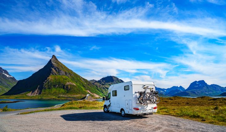 Motorhome bikes attached to the back parked in a scenic location next to a lake surrounded by mountains with plenty of greenery covering them and a blue sky